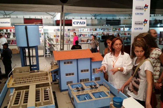 Na maqueta de uma ETA, servidora da Caema explica a visitantes do stand o processo de tratamento da água oferecida à população